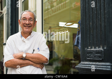 Senior hispanic coiffure à old fashion coiffure's shop, posing and looking at camera with arms crossed près de vitrine Banque D'Images