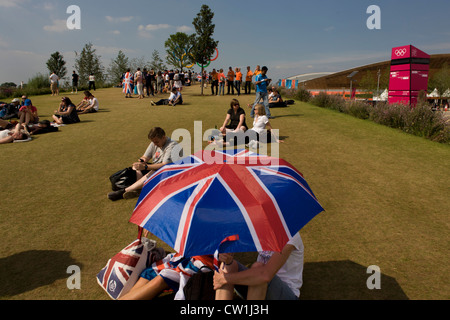 Brits profitez d'un été chaud pour regarder la couverture en direct d'un grand écran de télévision dans le Parc olympique au cours de l'Jeux olympiques de 2012 à Londres. En vertu de l'Union jack parasols ils s'asseoir sur l'herbe verte situé sur une colline où la loi sur les anneaux olympiques géant comme toile de fond pour les photos de famille. Ce terrain a été transformé pour devenir un 2,5 Km2 complexe sportif, une fois que les entreprises industrielles et maintenant le lieu de huit salles dont l'arène principale, centre aquatique et le vélodrome et le Village Olympique des athlètes. Après les Jeux Olympiques, le parc est d'être connu sous le nom de Queen Elizabeth Olympic Park. (Plus le sous-titrage dans Description..) Banque D'Images