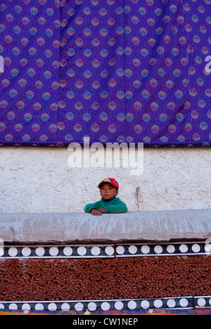 Jeune garçon dans une casquette de baseball rouge sur un balcon avec une thangka de soie en arrière de lui à Hemis Monastery, Ladakh, Inde Banque D'Images