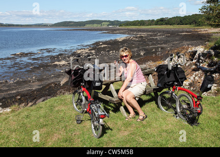 Mature Woman sitting on bench avec Brompton pliant près de Torrs Kirkcudbright Bay Point, Ecosse UK Banque D'Images