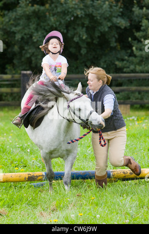 Une jeune fille sur un cheval blanc en cours d'avance sur un petit saut en bois dans un enclos d'herbe dans la campagne anglaise en été Banque D'Images