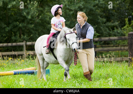 Une jeune fille sur un cheval blanc en cours d'avance sur un petit saut en bois dans un enclos d'herbe dans la campagne anglaise en été Banque D'Images