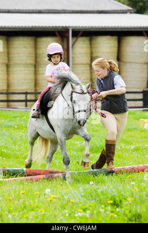 Une jeune fille sur un cheval blanc en cours d'avance sur un petit saut en bois dans un enclos d'herbe dans la campagne anglaise en été Banque D'Images