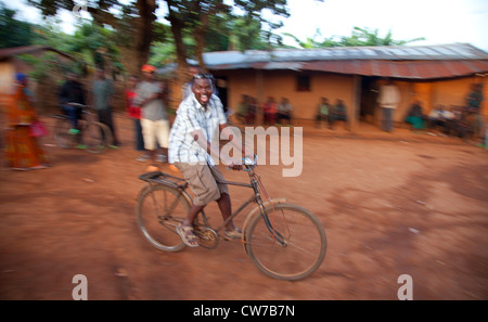 Homme ivre rire follement alors que rouler à vélo à travers un village de maisons en briques de boue, au Burundi, à proximité de Parc National de la Ruvubu, Cankuzo Banque D'Images