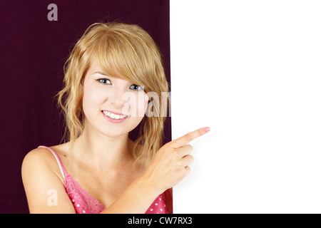 Mignon et sympathique jeune adolescent blond girl holding et en montrant un signe blanc vide, d'un placard ou d'affichage. Banque D'Images