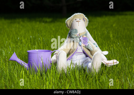 Lapin jouet mignon dans des vêtements assis sur l'herbe avec le peut, à l'extérieur Banque D'Images