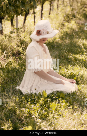 Une femme dans une robe victorienne blanche assise sur l'herbe entre les vignes Banque D'Images