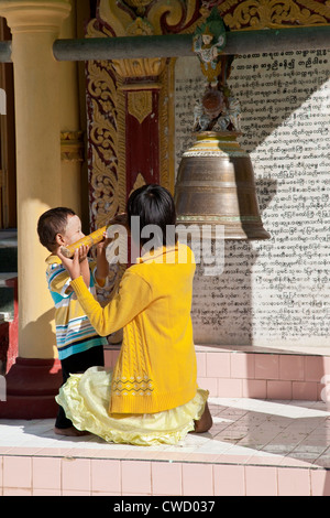 Le Myanmar, Birmanie. Une grande sœur enseigne son frère de grève la cloche du temple, à conduire des mauvais esprits de la maison. Banque D'Images