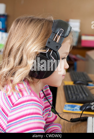 5 ans fille blanche est assis à un bureau et utilise des écouteurs pour écouter programme informatique Banque D'Images