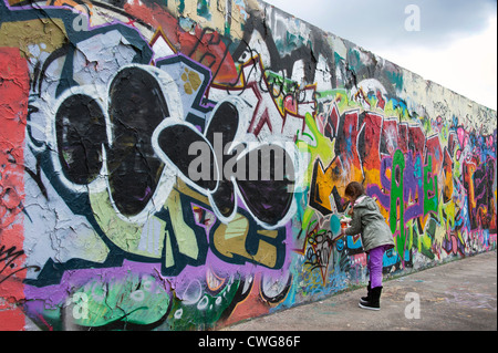 Une jeune fille ajoute sa note au dessus d'autres graffitis sur une partie du mur de Berlin, Berlin, Allemagne. Banque D'Images