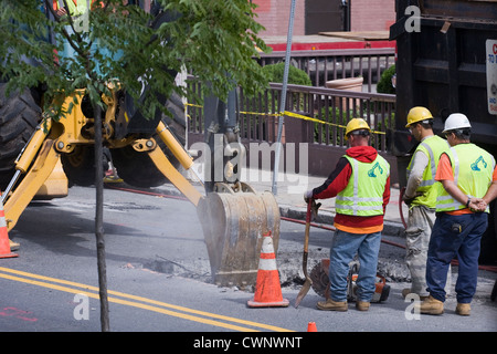 Trois travailleurs de la construction regarder une pelleteuse creuser un trou dans une chaussée. Banque D'Images