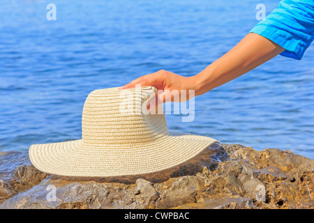 Woman's hand holding a hat à la plage contre le bleu de la mer en arrière-plan Banque D'Images