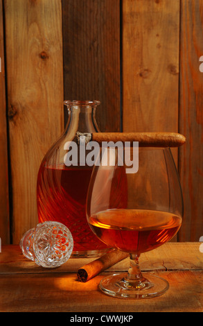 Verre de cognac en face d'une carafe en cristal sur un cadre en bois de style rustique. Deux cigares un allumé et posé sur le haut de la vitre. Banque D'Images