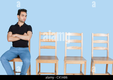 Portrait d'un jeune homme assis à côté de chaises en bois vides sur fond bleu Banque D'Images