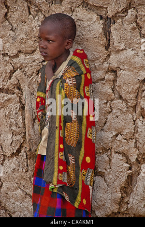 Enfant masaï, portant des vêtements traditionnels, debout contre un mur de boue dans un village de la Masai Mara, Kenya, Afrique Banque D'Images