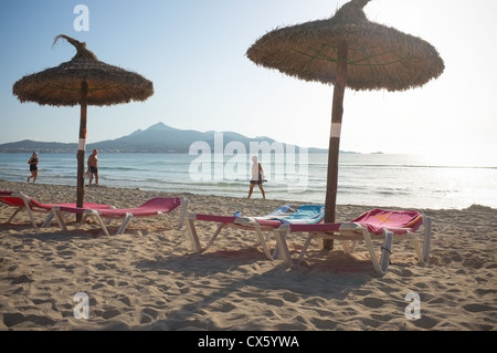 La plage d'Alcudia mallorca tôt le matin lorsque le soleil se lève avec les marcheurs et coureurs sur la plage vide à côté de chaises longues Banque D'Images