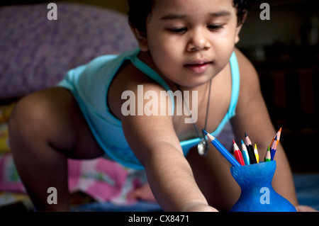 Une jeune Indienne joue avec les crayons de couleur conservés dans un petit vase bleu assise sur son lit. Banque D'Images