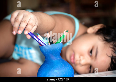 Une jeune Indienne joue avec les crayons de couleur conservés dans un petit vase bleu portant sur son lit. Banque D'Images
