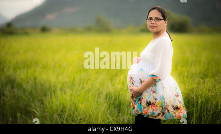 Belle asiatique femme portant des lunettes et affectueusement tenant son ventre enceinte dans un champ de riz nouvellement plantés Banque D'Images