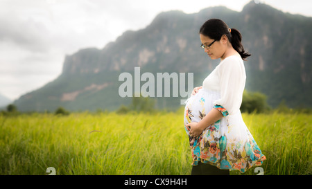 Belle asiatique femme portant des lunettes et affectueusement tenant son ventre enceinte dans un champ de riz nouvellement plantés Banque D'Images