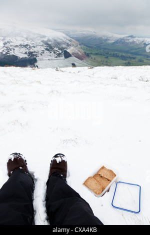 Randonneur avec des sandwichs emballés dans une boîte à lunch au sommet d'une colline dans la neige de l'hiver, Peak District, Derbyshire, Angleterre, RU Banque D'Images