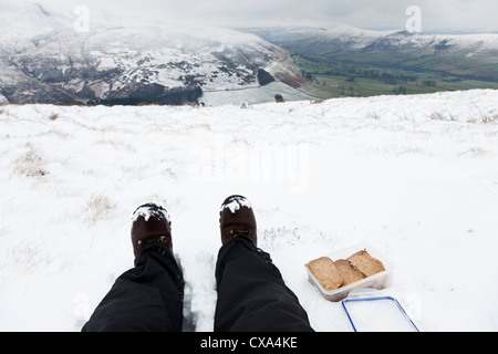 Walker de manger un panier-repas de sandwichs en restant assis dans la campagne de la neige durant l'hiver, Peak District, Derbyshire, Angleterre, RU Banque D'Images