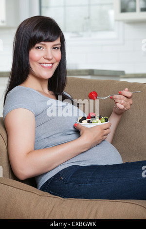 Portrait of pregnant woman eating fruit salad, assis dans le canapé Banque D'Images