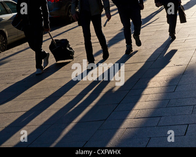 Les banlieusards de marcher sur la chaussée de la ville avec de longues ombres projetées par le soleil de fin d'après-midi Banque D'Images