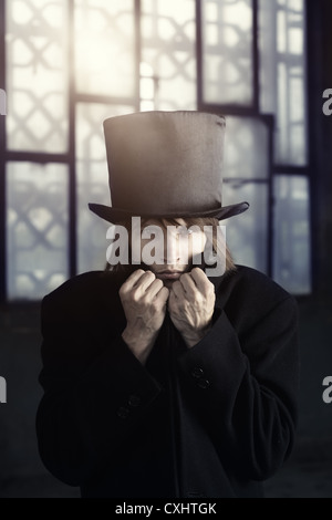 Seul l'homme à l'intérieur dans le manteau noir et un chapeau. Grande fenêtre à l'arrière-plan avec la lumière du soleil Banque D'Images