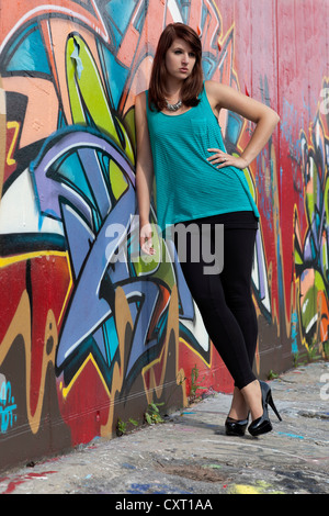 Jeune femme portant un haut turquoise, guêtres noires et de hauts talons en face de mur de graffiti Banque D'Images