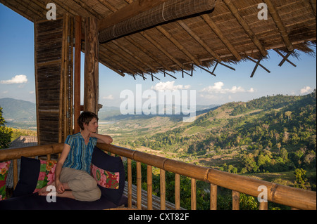 Femme assise sur un porche ou une véranda donnant sur le paysage, bamboo hut, Lanjia Lodge, dans le Nord de la Thaïlande, la Thaïlande, l'Asie Banque D'Images