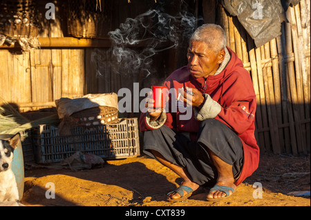 Man smoking a cigarette et boire le thé en face d'une case en paille, village de la tribu Akha hill, minorité ethnique Banque D'Images