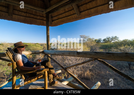 Femme avec un chapeau assis sur une chaise tenant un verre de champagne, plate-forme panoramique, zoo privé, Limpopo, Afrique du Sud, l'Afrique Banque D'Images
