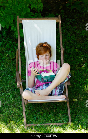 Enfant, garçon, 11 ans, la lecture d'une maison de vacances passionnant livre tout en se reposant dans une chaise longue dans le jardin Banque D'Images