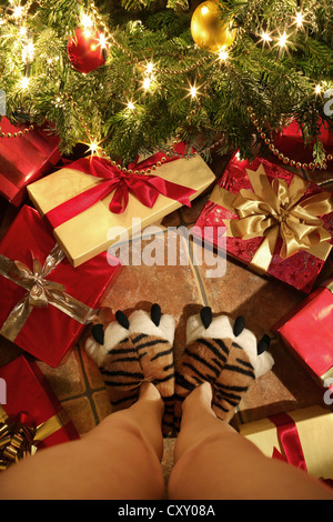 Personne avec des chaussons en forme de pattes de tigre debout devant des cadeaux de Noël sous un arbre de Noël Banque D'Images
