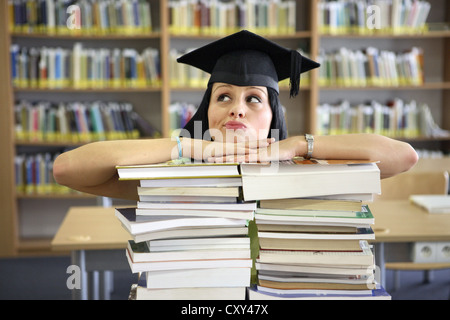 Sceptique quant à la recherche étudiante portant une graduation cap derrière des piles de livres dans une bibliothèque universitaire Banque D'Images