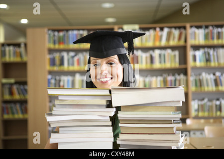 Smiling female student portant une graduation cap derrière des piles de livres dans une bibliothèque universitaire Banque D'Images