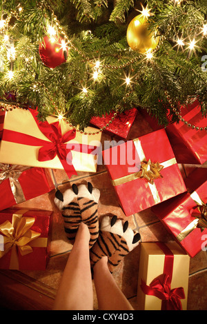 Personne avec des chaussons en forme de pattes de tigre debout devant des cadeaux de Noël sous un arbre de Noël Banque D'Images