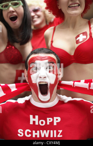 Jeune homme hurlant, fan de football avec un visage peint, drapeau national suisse, portant un t-shirt rouge, lettrage "Hopp Schwiiz" Banque D'Images