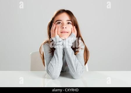 Belle petite fille assise derrière un bureau et de penser, sur un arrière-plan gris Banque D'Images