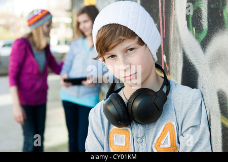 Boy wearing headphones debout devant un mur de graffiti, avec deux filles derrière lui Banque D'Images