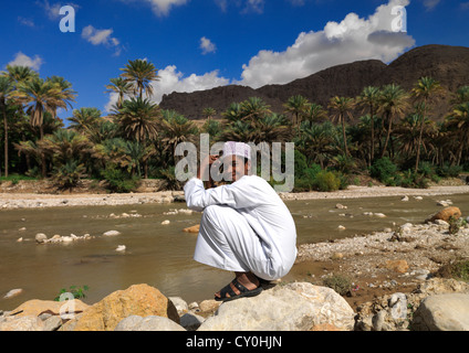 Garçon accroupi sur le rocher à côté de la rivière avec un fond de Palmiers, Wadi Bani Khalid, Oman Banque D'Images