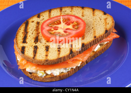 Sandwich au saumon fumé et salade russe décoré d'une tranche de tomate servi sur une plaque bleue sur un fond orange. Banque D'Images