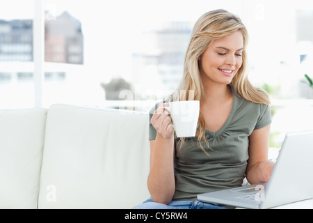 Woman smiling comme elle tient sa tasse et utilise son ordinateur portable Banque D'Images