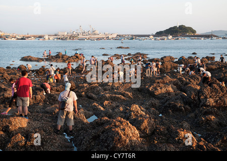 Les vacanciers recherchez les rochers piscines à Qingdao alors qu'un destroyer de la retraite de la Shanghai musée naval se trouve dans l'arrière-plan Banque D'Images