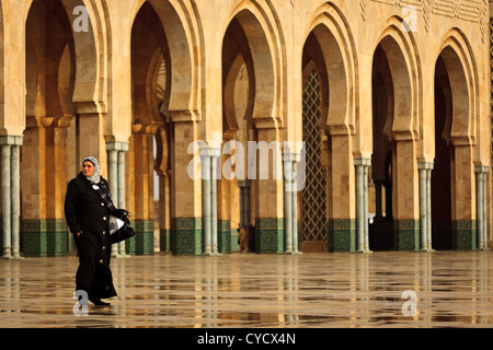 Femme dans un long manteau noir et foulard marcher devant orné de voûtes de mosquée Hassan II, Casablanca, Maroc. Banque D'Images