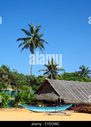 Une scène de plage tropicale avec un outrigger canoe et de huttes au toit de chaume avec des palmiers sous un ciel bleu clair Banque D'Images