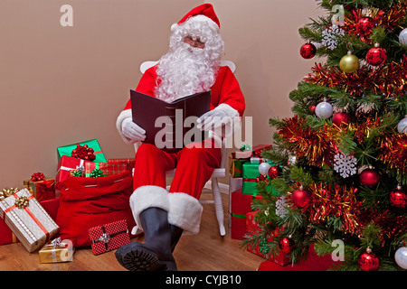 Santa Claus était assis dans une chaise à bascule la lecture de la 'liste' Gentil ou méchant entouré de gift wrapped presents. Banque D'Images