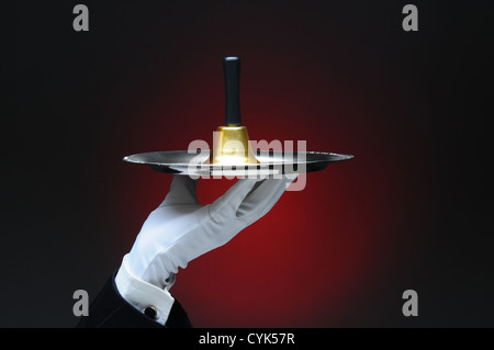 Closeup of a white gloved hand holding bell un service sur un plateau d'argent format horizontal sur un fond rouge clair à foncé. Banque D'Images