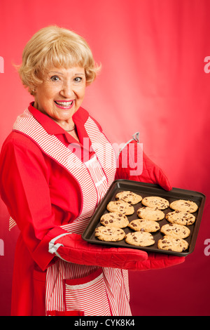 Grand-mère au foyer doux holding a tray of fresh biscuits aux brisures de chocolat. Photographié en face de fond rouge. Banque D'Images